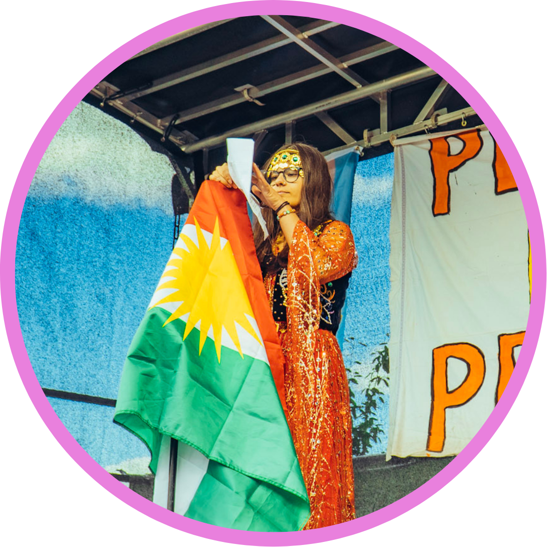 Elleonora har långt brunt hår, glasögon och en traditionell kurdisk röd klänning. Hen står på en scen och håller i ett manuspapper och en kurdisk flagga. Bakgrunden är en blå himmel.